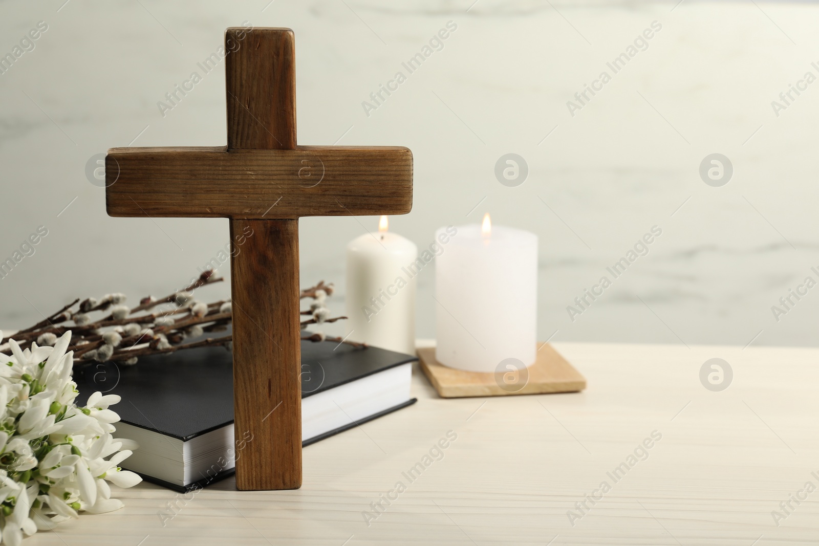 Photo of Wooden cross, Bible, church candles, willow branches and snowdrops on white table. Space for text