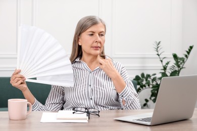 Photo of Menopause. Woman waving hand fan to cool herself during hot flash at wooden table indoors