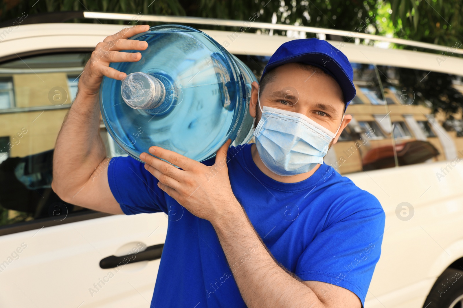 Photo of Courier in medical mask holding bottle of cooler water near car outdoors. Delivery during coronavirus quarantine