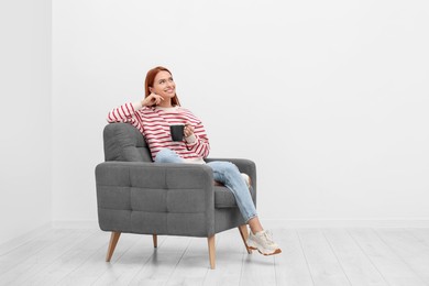Happy young woman with cup of drink sitting in armchair near white wall indoors. Space for text