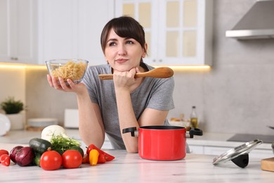 Photo of Pensive housewife with spoon and raw pasta at white marble table in kitchen. Cooking process