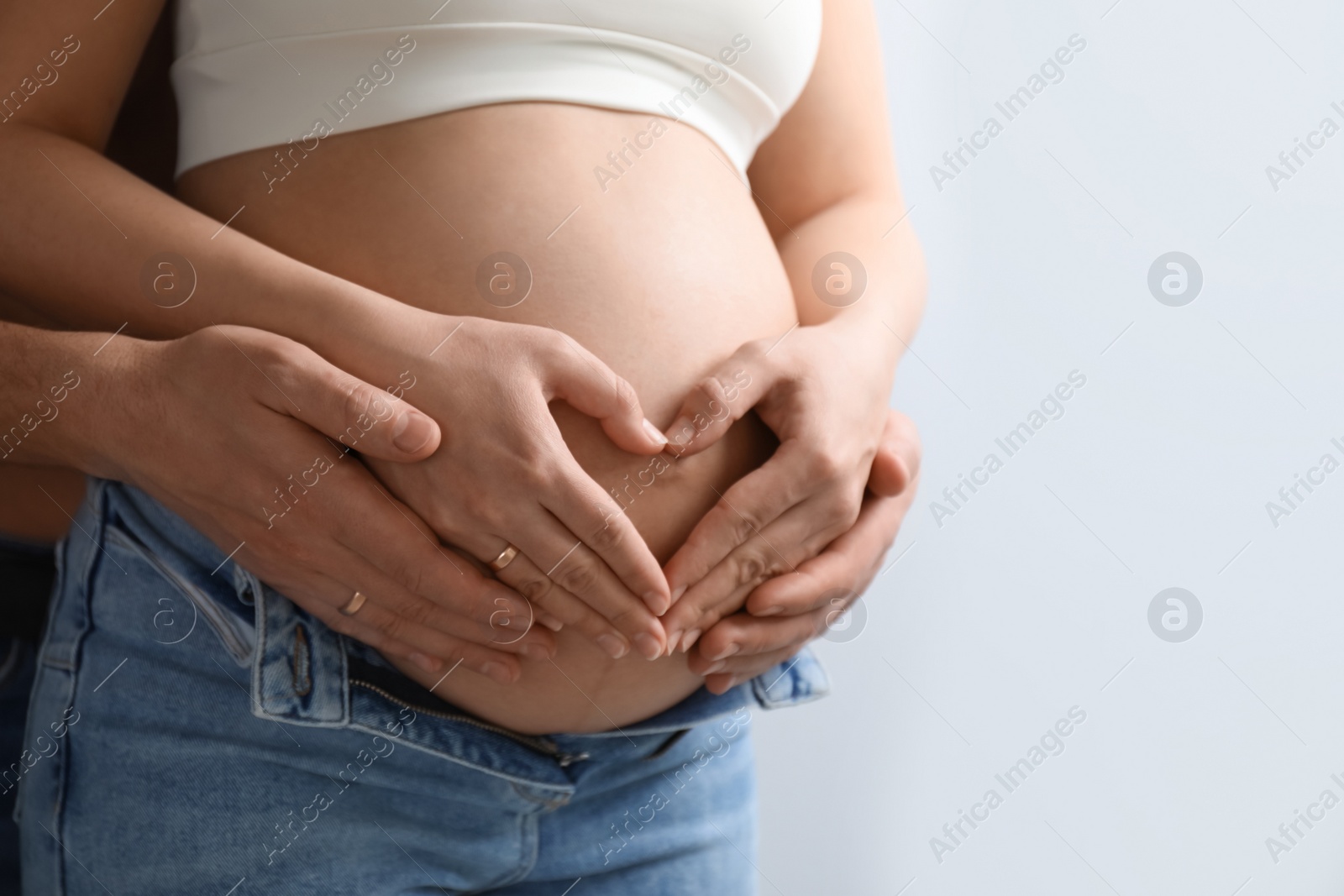 Photo of Pregnant young woman making heart with hands on belly and husband near her against light background, closeup