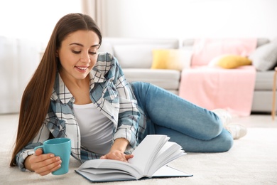 Photo of Beautiful young woman with cup of coffee reading book on floor at home. Space for text