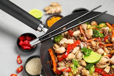 Photo of Stir-fry. Tasty noodles with meat in wok, chopsticks and ingredients on grey textured table