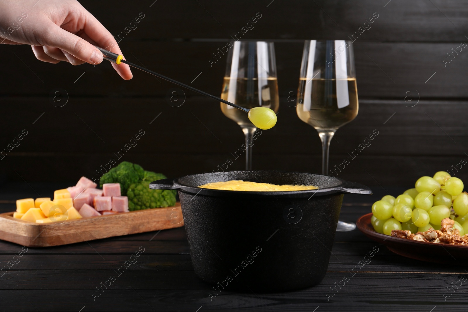 Photo of Woman dipping grape into fondue pot with melted cheese at black wooden table, closeup