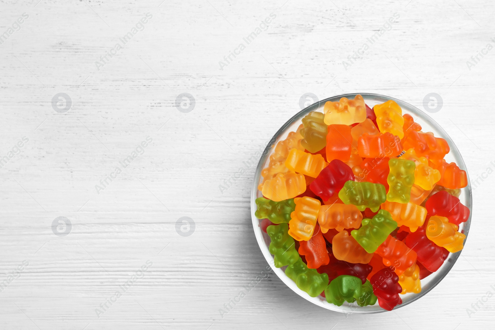 Photo of Bowl with delicious bright jelly bears on white wooden table, top view. Space for text