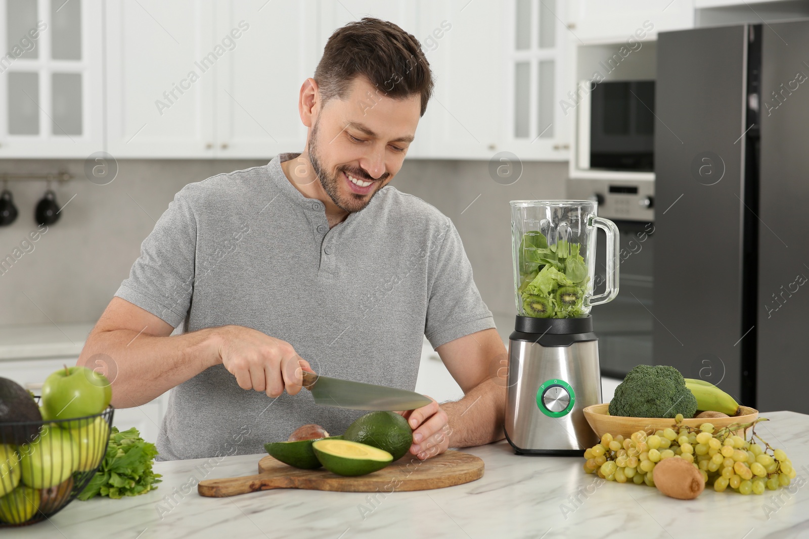 Photo of Happy man cutting avocado for delicious smoothie at white marble table in kitchen