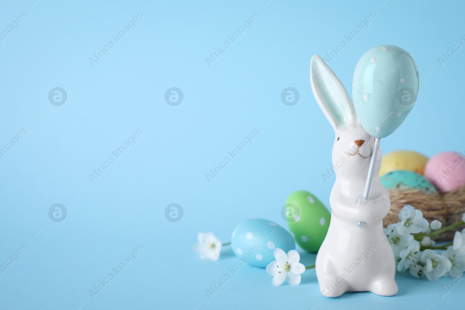 Photo of Easter bunny figure, flowers and dyed eggs on light blue background. Space for text