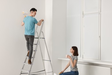 Photo of Young man painting wall on stepladder while woman drinking coffee indoors. Room renovation