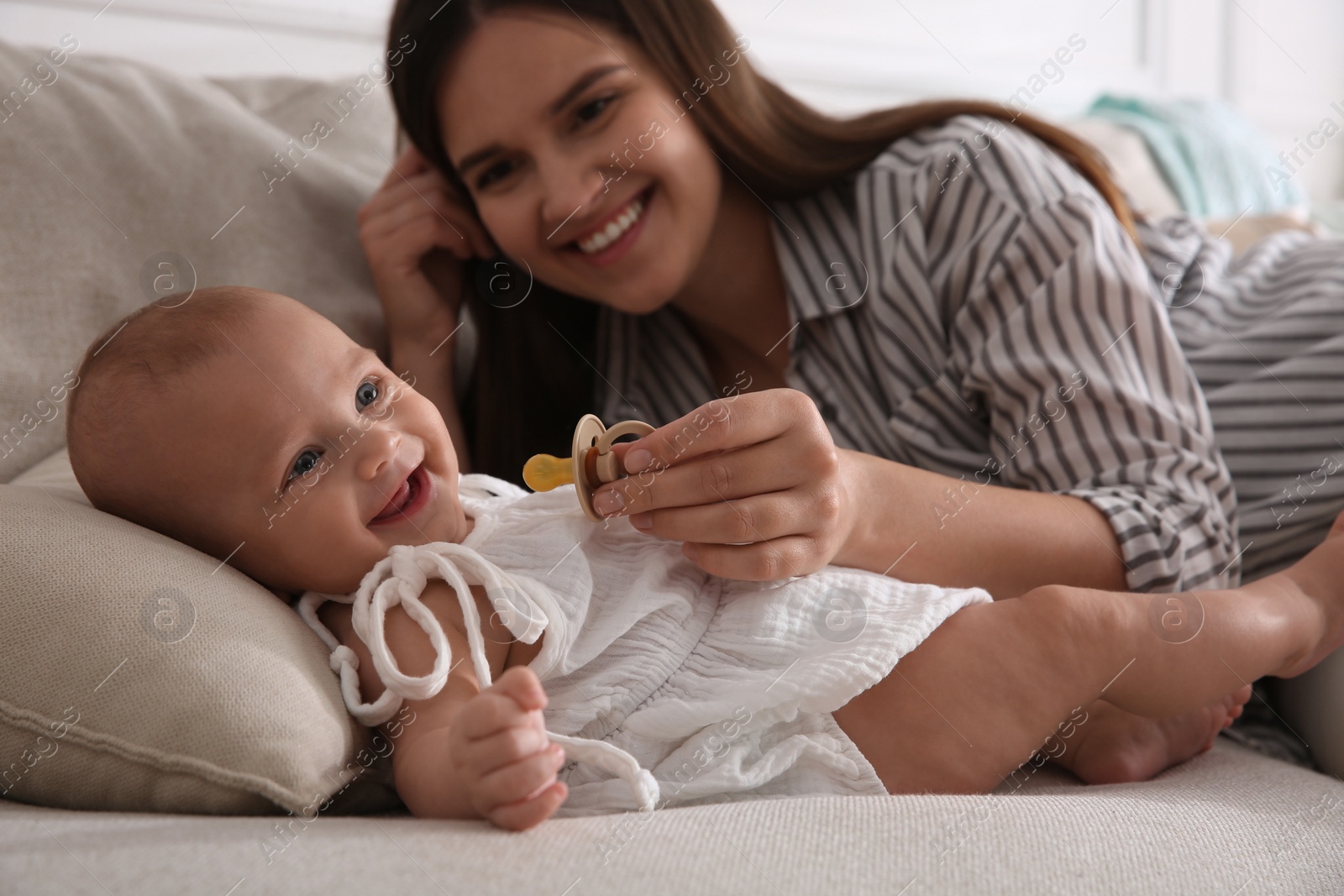 Photo of Happy mother giving pacifier to her cute little baby on sofa at home