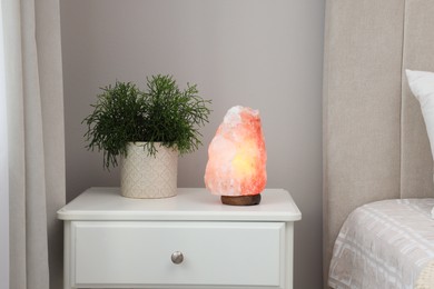 Beautiful Himalayan salt lamp and green houseplant on white stand in bedroom