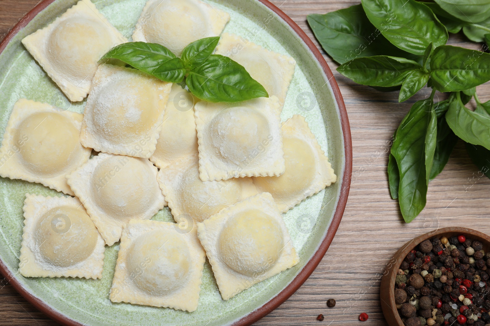 Photo of Uncooked ravioli, basil and peppercorns on wooden table, flat lay