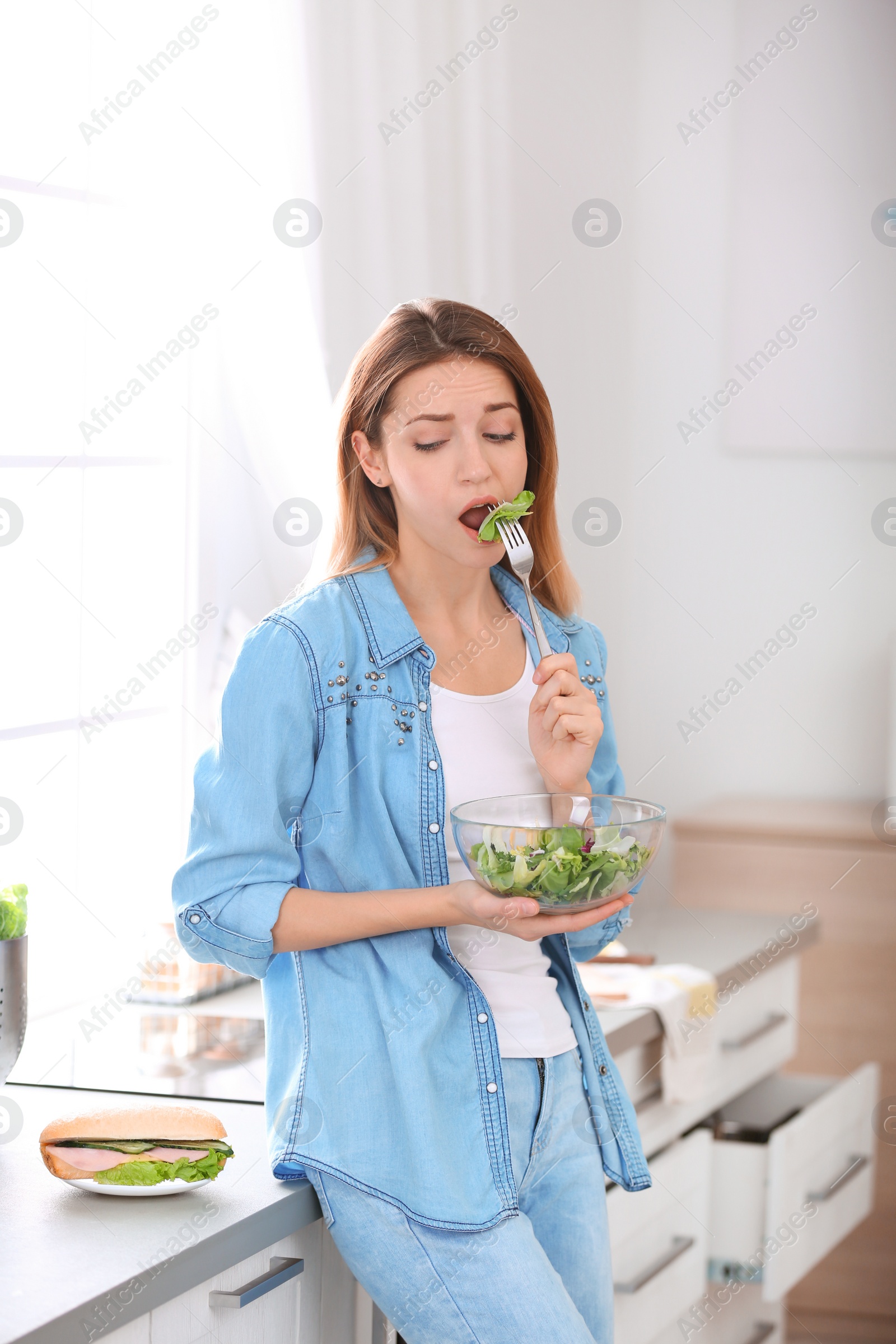 Photo of Emotional young woman eating salad instead of sandwich in kitchen. Healthy diet