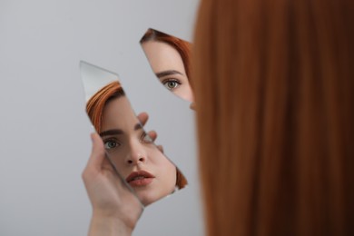 Photo of Young woman looking at herself in shard of broken mirror on light grey background, closeup