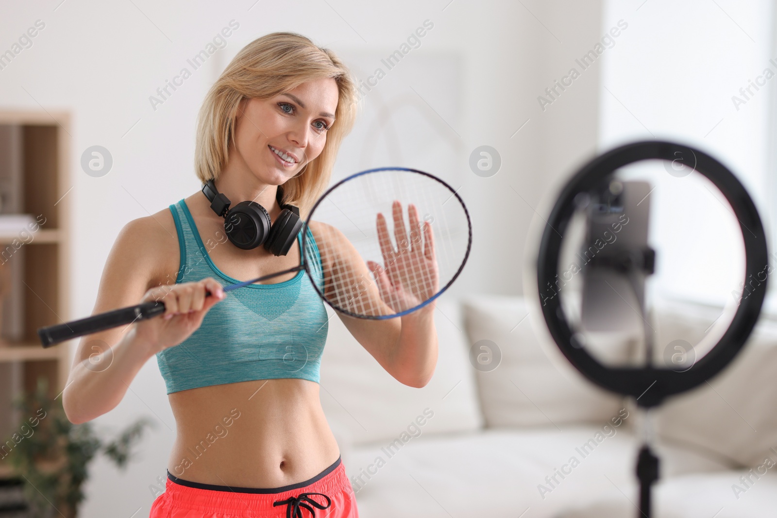 Photo of Smiling sports blogger holding badminton racket while streaming online fitness lesson with smartphone at home