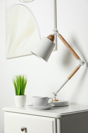 Photo of Stylish lamp, houseplant and cup of drink on white side table in light room. Interior design