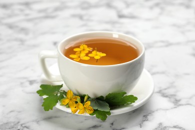 Photo of Cup of aromatic celandine tea and flowers on white marble table, closeup