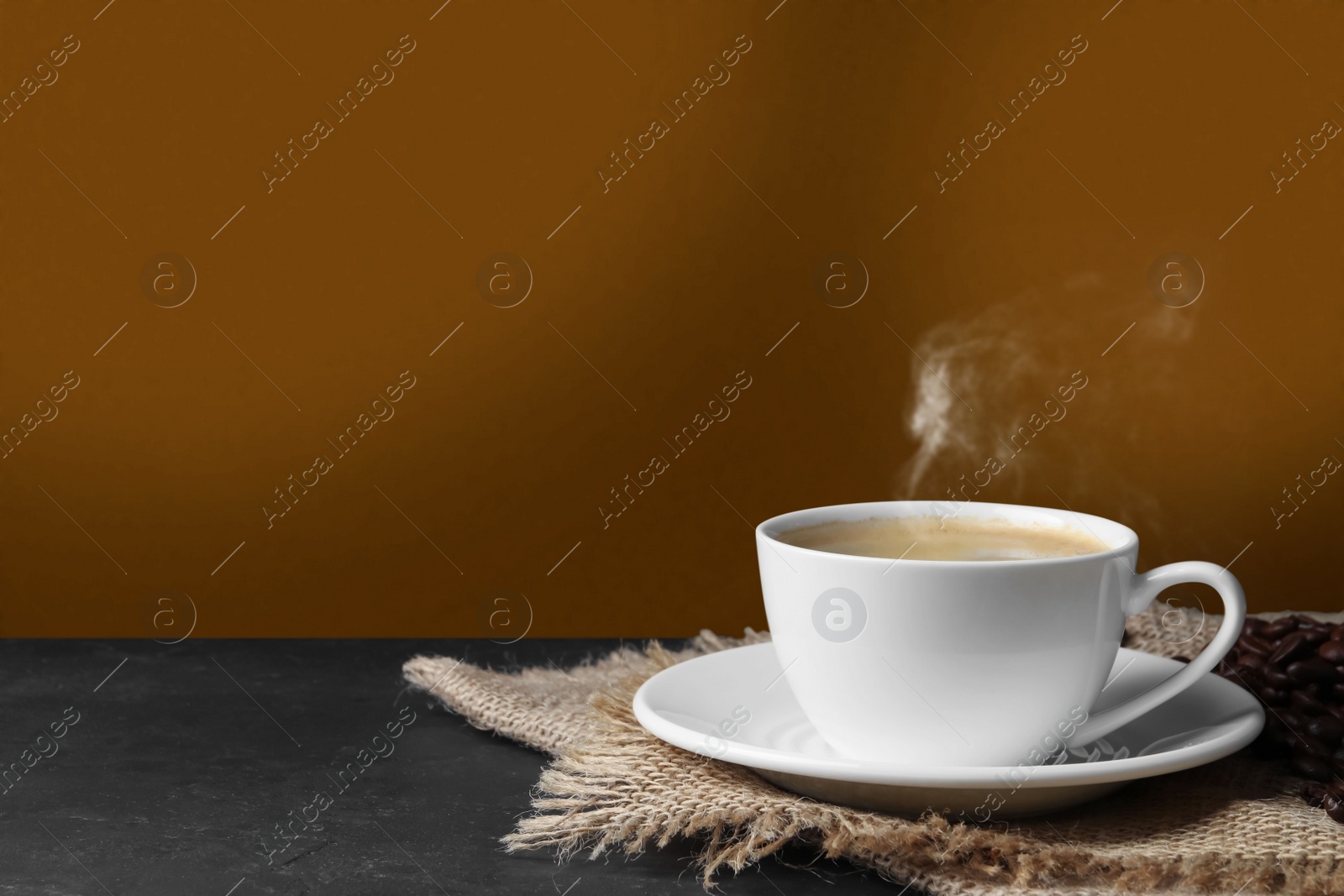 Image of Cup of hot aromatic coffee and roasted beans on black table against brown background. Space for text