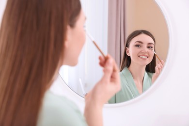 Photo of Beautiful young woman applying eyeshadow with brush near mirror indoors