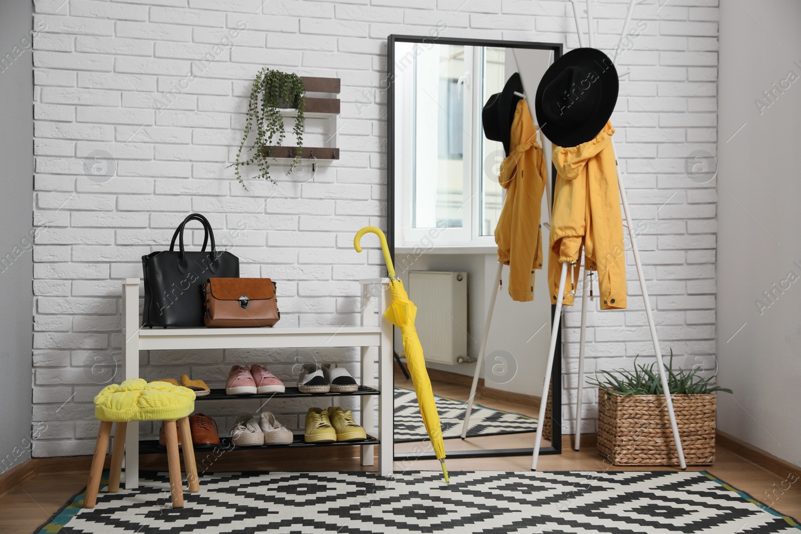 Photo of Stylish hallway interior with coat rack, shoe storage bench and mirror near white brick wall