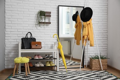 Stylish hallway interior with coat rack, shoe storage bench and mirror near white brick wall