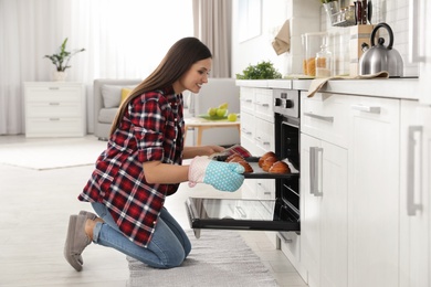 Beautiful woman taking out tray of baked buns from oven in kitchen