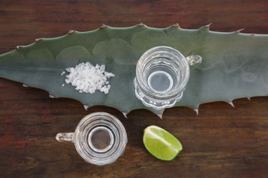 Photo of Mexican tequila shots, salt, lime slice and green leaf on wooden table, flat lay. Drink made of agava