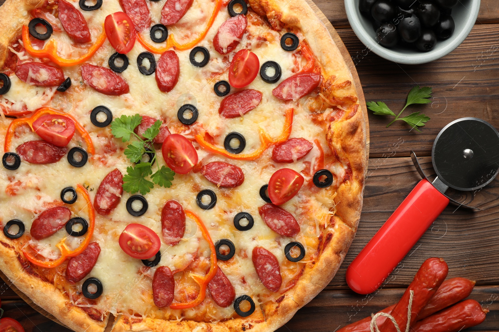 Photo of Tasty pizza with dry smoked sausages, tomato, olives and cutter on wooden table, top view