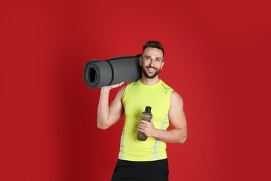 Handsome man with yoga mat and bottle of water on red background