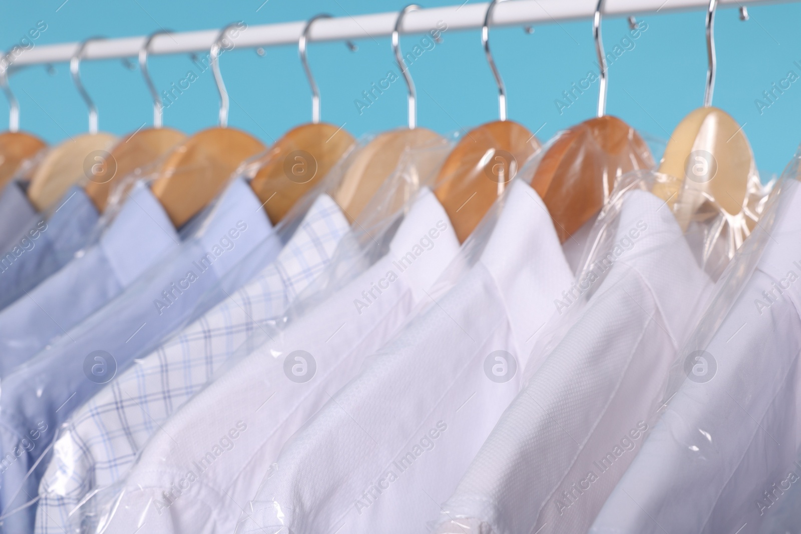 Photo of Dry-cleaning service. Many different clothes in plastic bags hanging on rack against light blue background, closeup