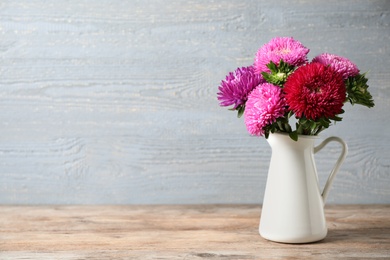 Photo of Jug with beautiful aster flowers on table against wooden background. Space for text