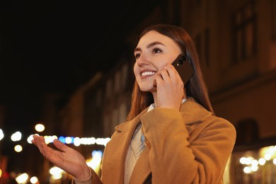 Smiling woman talking by smartphone on night city street. Space for text