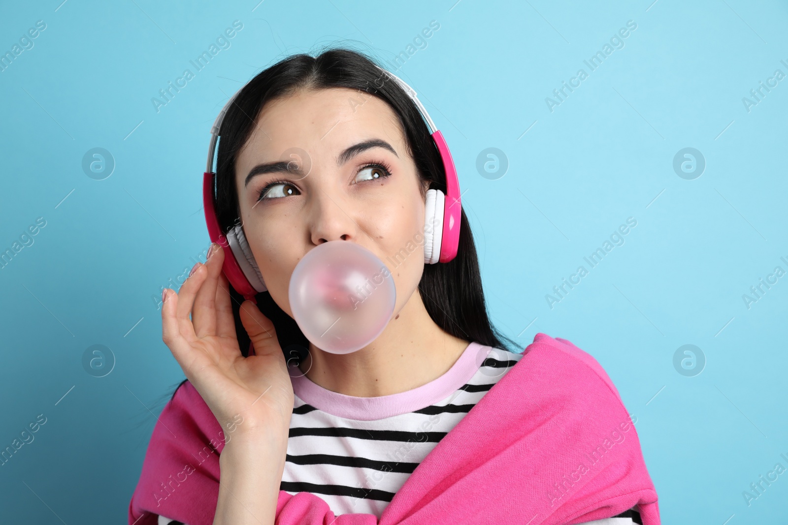 Photo of Fashionable young woman with headphones blowing bubblegum on light blue background
