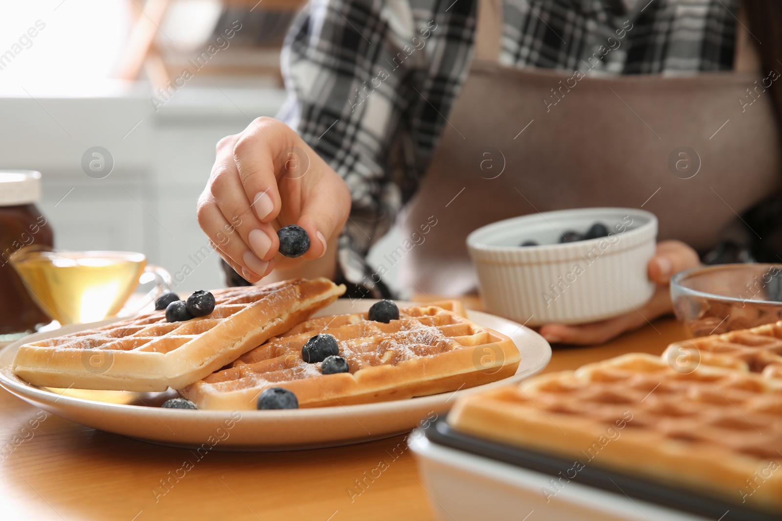 Photo of Woman decorating delicious Belgian waffles with blueberries at wooden table in kitchen, closeup