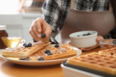 Photo of Woman decorating delicious Belgian waffles with blueberries at wooden table in kitchen, closeup