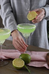 Photo of Woman squeezing lime juice into glass with delicious Margarita cocktail at wooden table, closeup