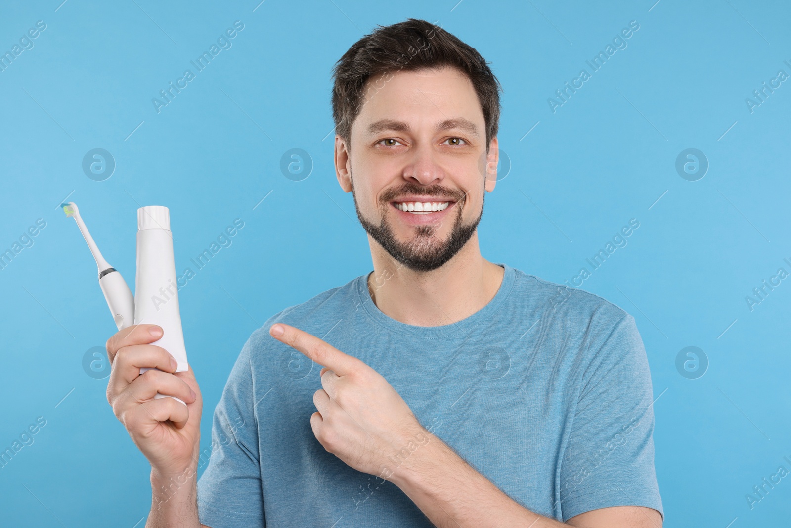 Photo of Happy man holding electric toothbrush and tube of toothpaste on light blue background