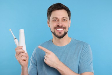 Happy man holding electric toothbrush and tube of toothpaste on light blue background