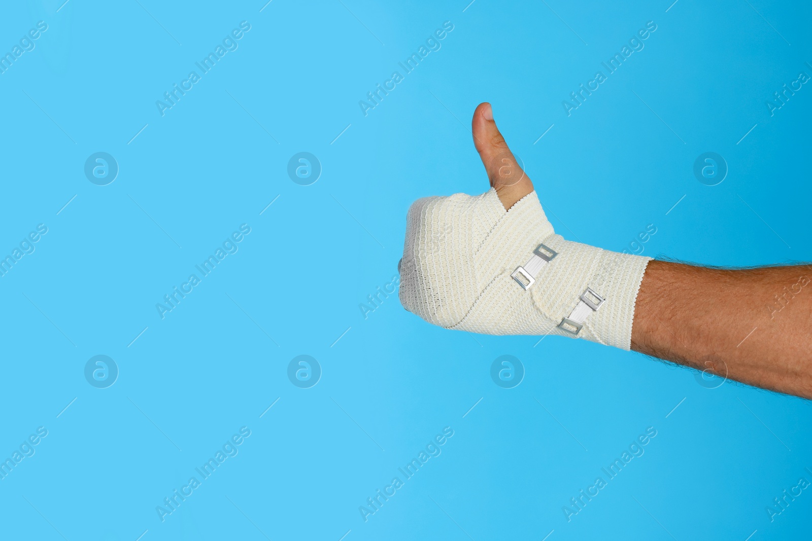 Photo of Man with hand wrapped in medical bandage showing thumbs up on light blue background, closeup. Space for text