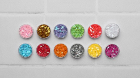 Photo of Many different colorful sequins in containers on white tiled background, top view