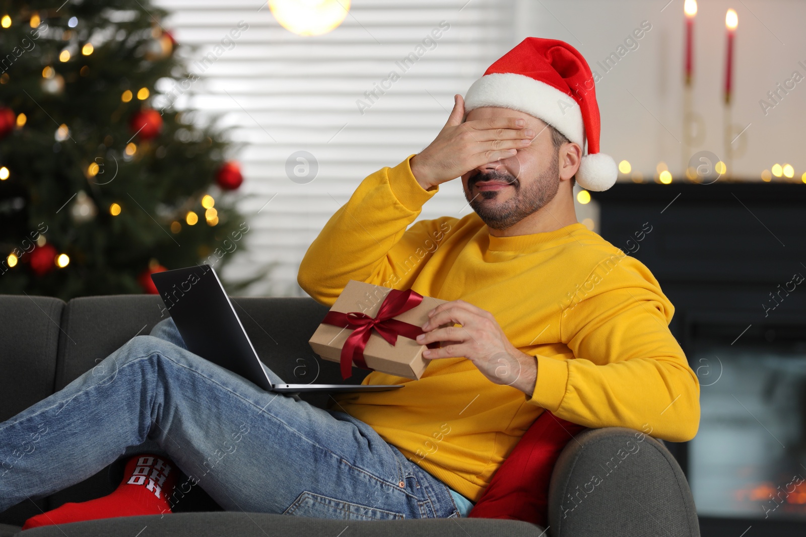 Photo of Celebrating Christmas online with exchanged by mail presents. Man in Santa hat covering eyes before opening gift box during video call on laptop at home