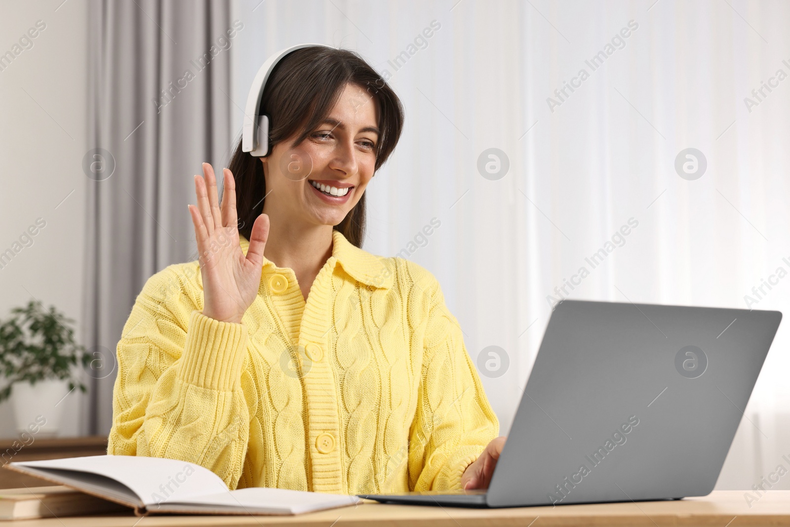 Photo of Happy woman waving hello during video chat via laptop at table indoors