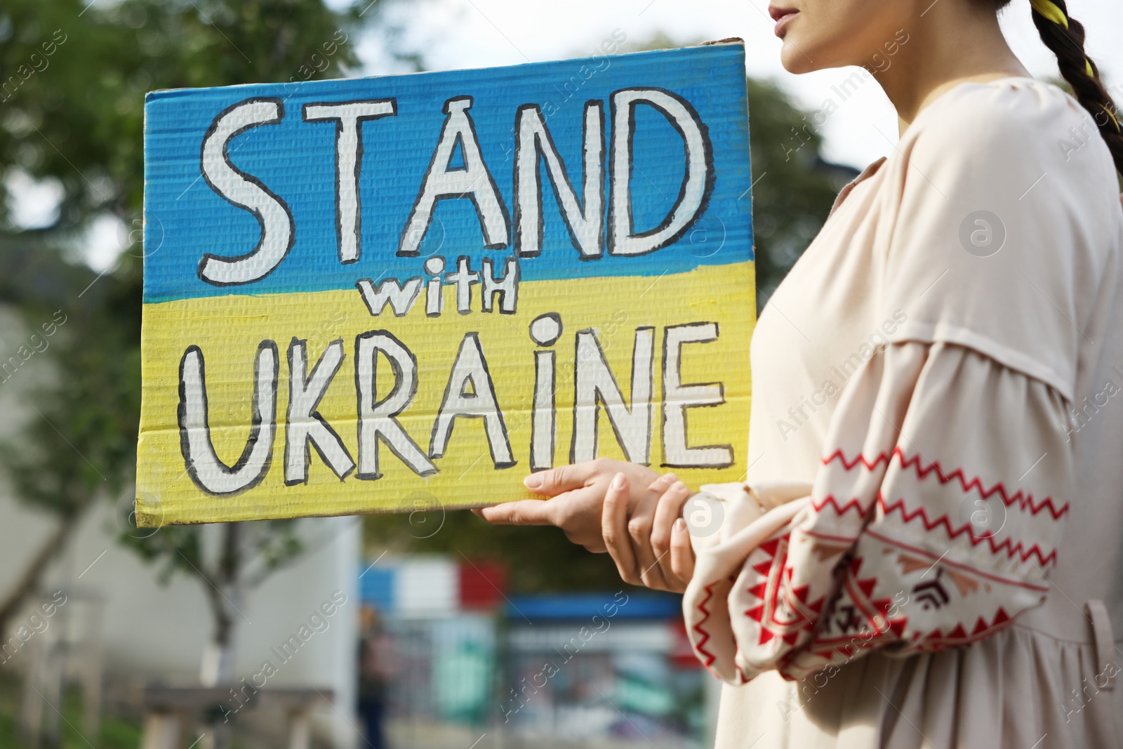 Photo of Young woman holding poster in colors of national flag and words Stand with Ukraine outdoors, closeup