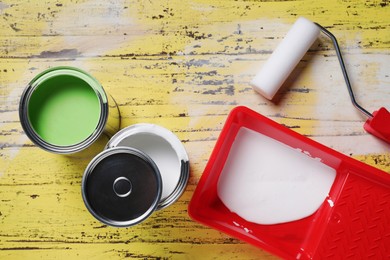 Cans of paints, roller and tray on yellow wooden table, flat lay