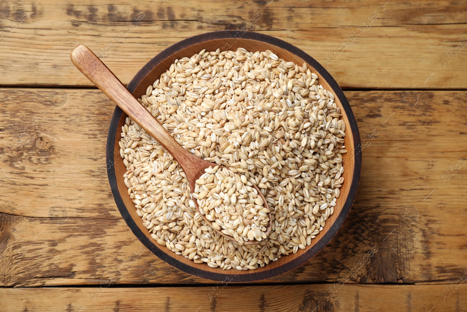 Photo of Dry pearl barley in bowl and spoon on wooden table, top view