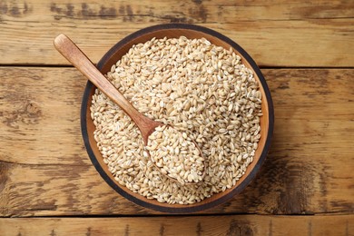 Dry pearl barley in bowl and spoon on wooden table, top view