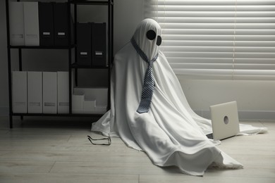 Photo of Overworked ghost. Man in white sheet with laptop on floor in office