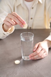 Photo of Woman putting effervescent pill into glass of water at grey table, closeup
