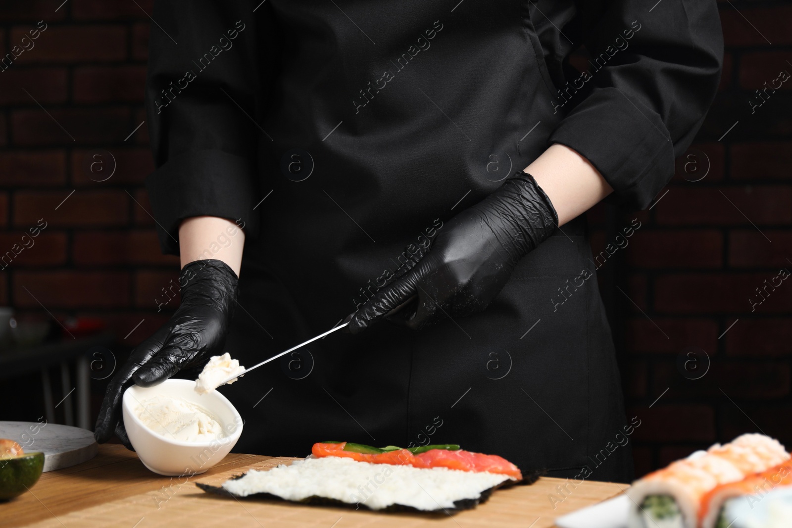 Photo of Chef in gloves putting cream cheese onto unwrapped sushi roll at wooden table, closeup