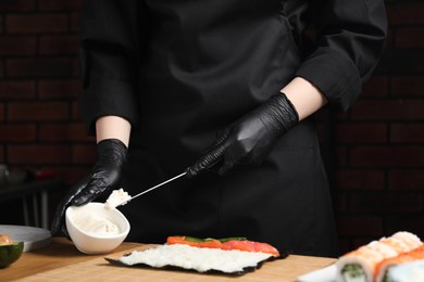 Chef in gloves putting cream cheese onto unwrapped sushi roll at wooden table, closeup